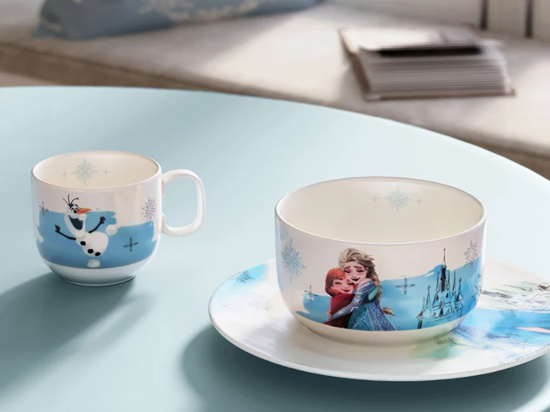Un juego de tazas y tazas de la colección Disney Frozen sobre una mesa azul. Un juego de tazas y tazas con personajes de la película Frozen, colocado sobre una mesa azul junto a una ventana.