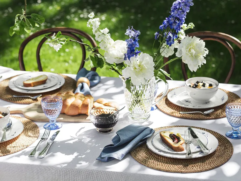 Outdoor table setting with flowers, blue glasses, bread and Old Luxembourg plates with food on woven placemats in a garden.