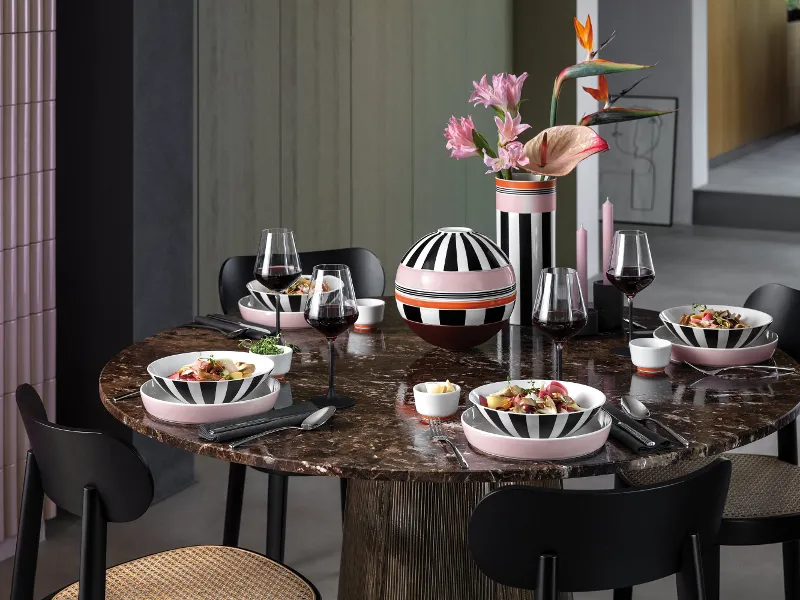 Ein runder Marmortisch mit vier Gedecken, gestreiftem Memphis Geschirr, Weingläsern und einer Vase mit rosa Blumen.