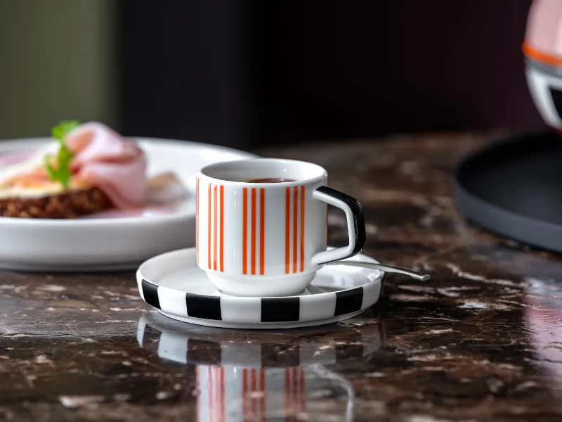 Eine gestreifte Memphis Espressotasse auf einer Untertasse steht auf einem Marmortisch, neben einem Memphis Teller

