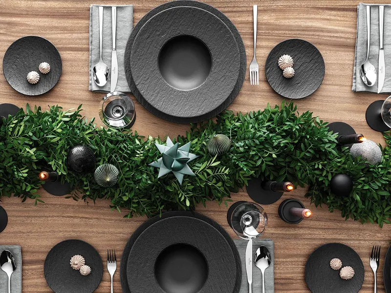 A top view of a Christmas dining table setting with black plates from the Villeroy & Boch Manufacture range together with Piemont cutlery. It is also decorated with a centrepiece of green foliage and candles on a wooden surface.