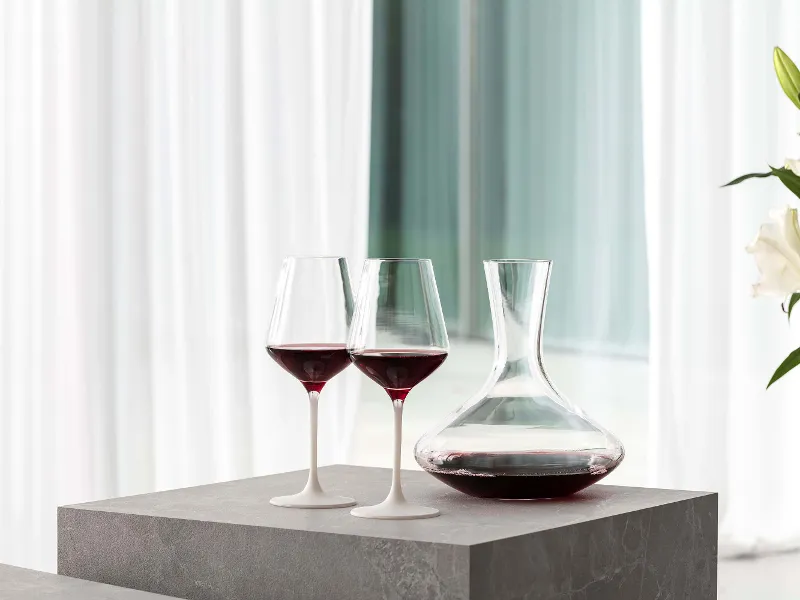 Two filled Manufacture red wine glasses on a table next to an elegant Purismo wine decanter.