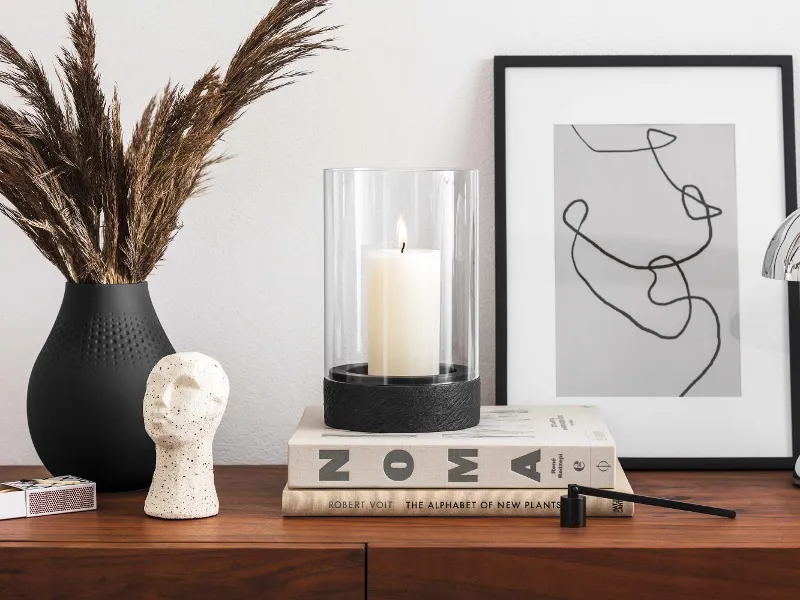 A modern highboard with decorative objects, including a vase with dried plants, a black Manufacture hurricane lamp, a sculpture, stacked books and a framed abstract artwork.