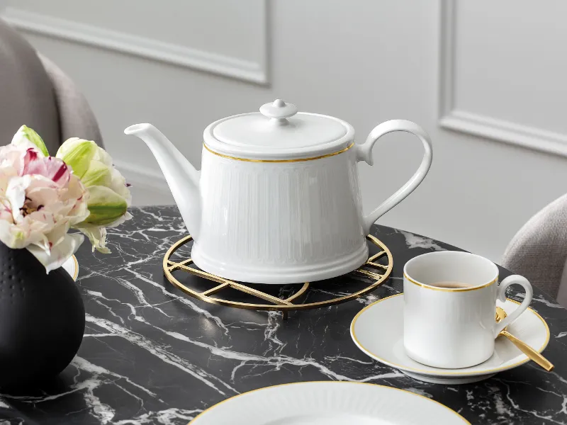 A white coffee pot with a gold rim on a stand on a marble table. Next to it is a matching teacup and saucer with a gold spoon and a black vase with a bouquet of flowers.