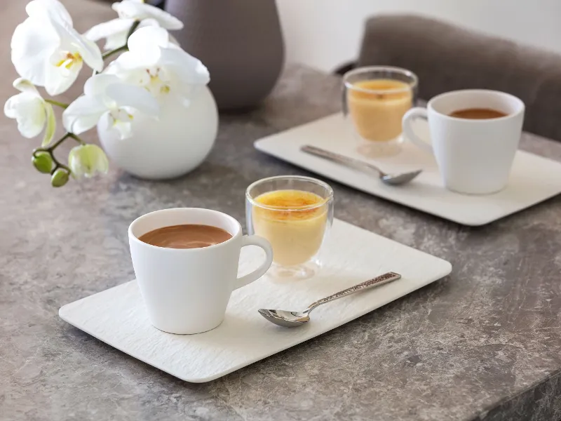 Zwei Artesano Espressotassen und zwei Desserts auf weißen rechteckigen Tellern mit Löffeln, auf einem Marmortisch neben einer weißen Orchidee von Villeroy & Boch.