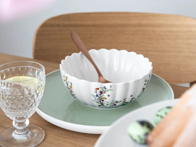 A decorative bowl with a floral pattern from the Spring Fantasy collection by Villeroy & Boch and a spoon on a green plate. A glass of water with lemon and a plate of assorted macarons are placed next to it on the table.