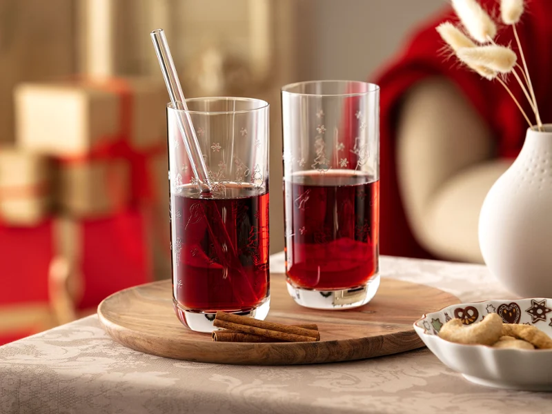 Toy's Delight longdrink glasses with festive red drink Two elegant Toy's Delight crystal longdrink glasses with delicate festive engravings, filled with a red holiday drink and a glass straw, set on a round wooden tray with cinnamon sticks in a cozy Christmas living room