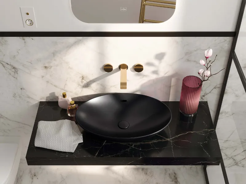 Schwarzes Aufsatzwaschbecken auf schwarzem Marmorwaschtisch mit goldenem Wasserhahn, Handtuch, Toilettenartikeln und rosa Vase mit Blumen.
