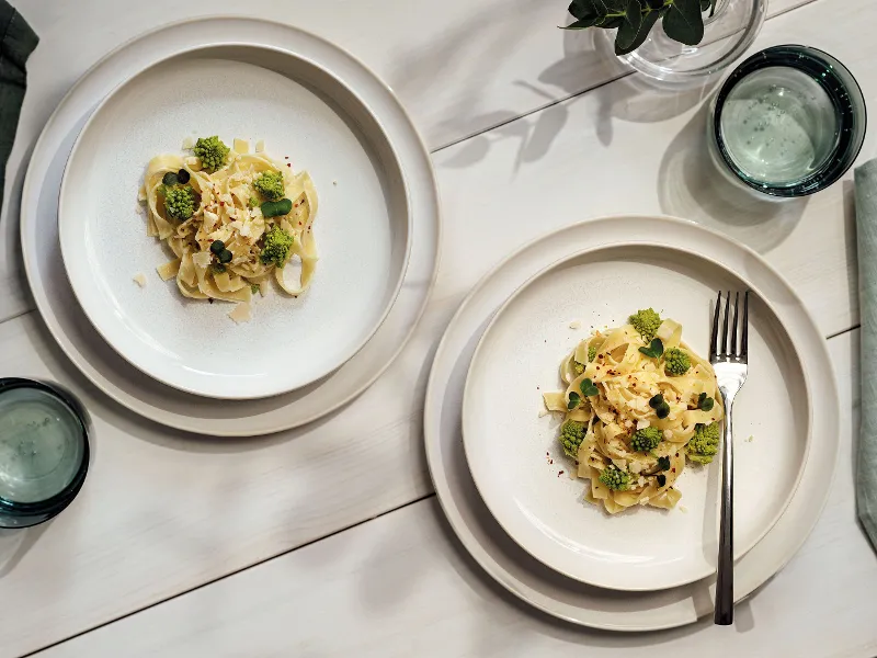 Two Crafted plates with broccoli and sauce on a white table, along with glasses of water and a small potted plant.