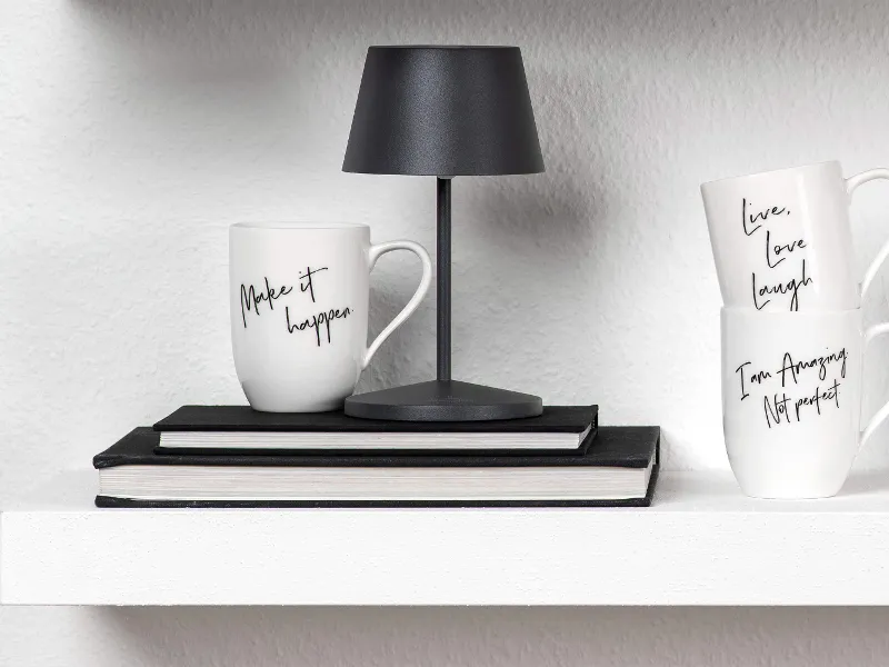 Tassen mit motivierenden Sprüchen, eine kleine schwarze Lampe und Bücher, angeordnet auf einem weißen Regal.
