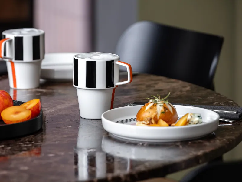 Memphis Kaffeetassen und ein Memphis Teller mit Essen und einem Stück Obst auf einem Marmortisch neben einem schwarzen Stuhl.