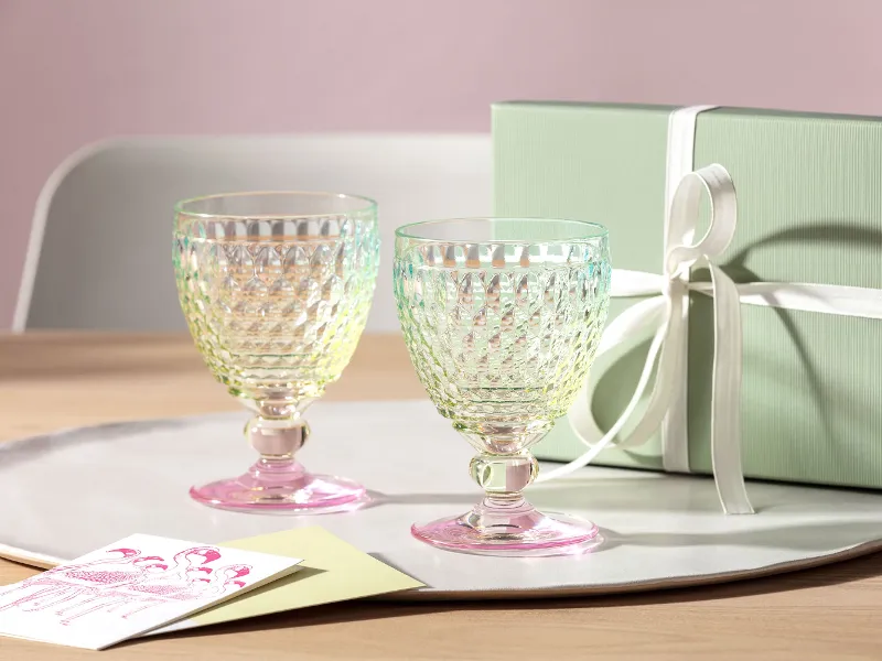 Two Boston glasses by Villeroy & Boch with a pink base are placed on a tray next to a green gift box with a white ribbon and a greeting card on a wooden table.