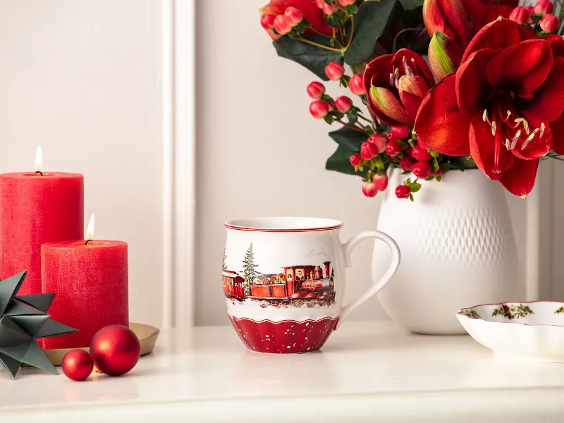 Red candles, a festive mug, red ornaments, and a vase with red flowers on a white surface.