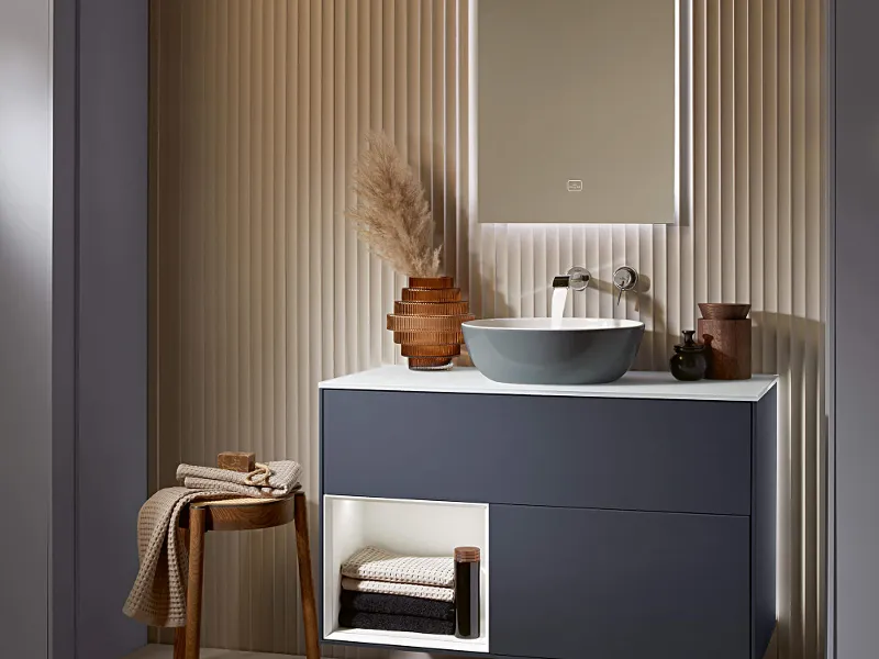 An Artis washbasin on a grey vanity unit and an illuminated mirror in a bathroom.