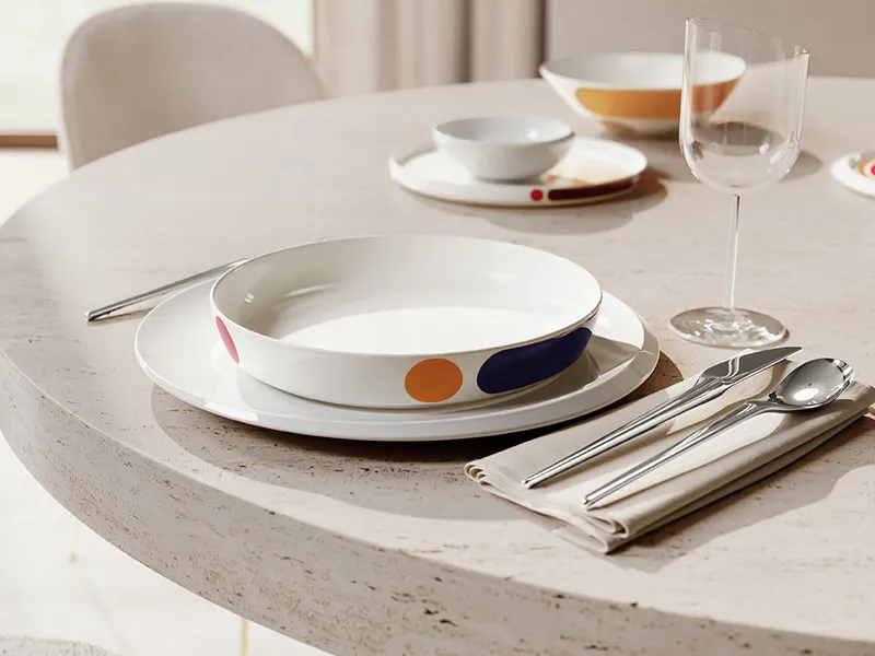 Tavola rotonda apparecchiata per un pasto, con piatto New Moon, ciotola La Boule Miami e posate New Moon su tovagliolo. Le stoviglie sono caratterizzate da motivi vivaci e colorati. Un calice da vino è accanto al tavolo.