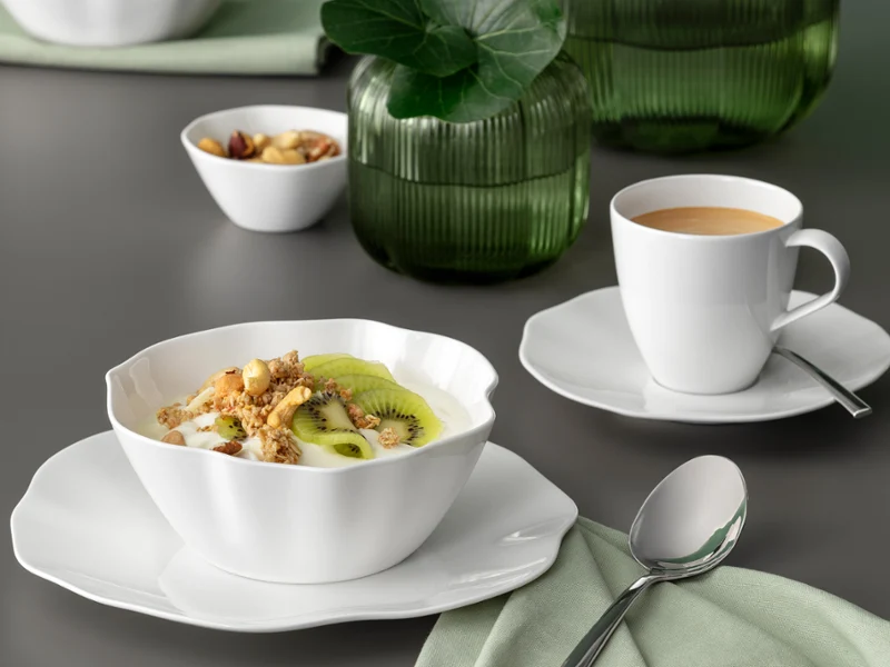A white Pura bowl with muesli and kiwi and a cup of coffee on a saucer A white Pura bowl with muesli and kiwi and a cup of coffee on a saucer on a grey table with a green napkin.