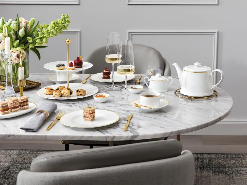 Ein für den Nachmittagstee gedeckter Marmortisch mit Desserts, Gebäck, Teeservice, Champagnergläsern und einem Blumenarrangement.