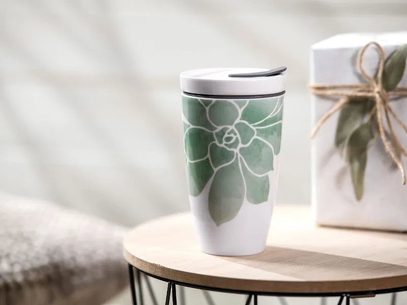 The Coffee ToGo mug from Villeroy & Boch, a ceramic travel mug with a green leaf design, stands on a wooden table next to a gift box tied together with string.