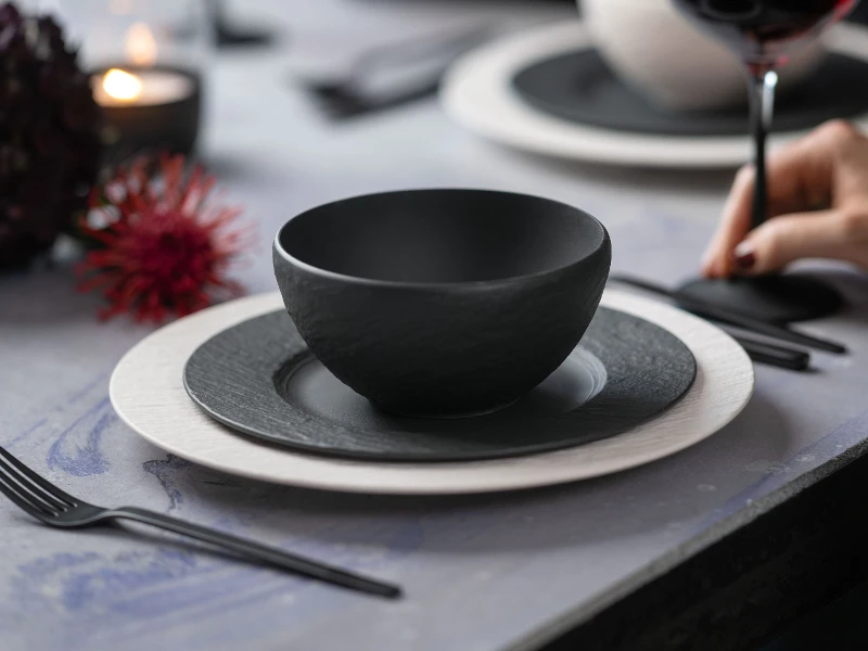 A table set with a black bowl, black and white plates, and black flatware. In the background, a hand holds a wine glass.