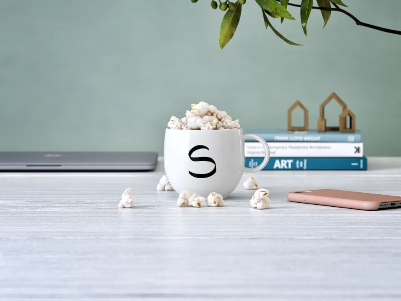 A Letters mug from Villeroy & Boch with the letter "s," filled with popcorn, on a desk with a laptop, books and a smartphone nearby.