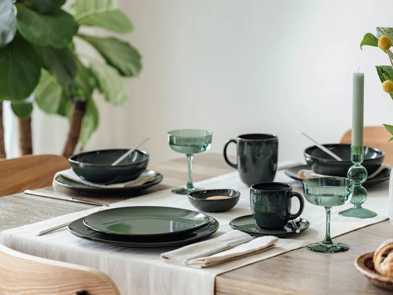 Ein Esstisch mit grünem Geschirr, Glaswaren, Besteck, einer Kerze und einer Vase mit gelben Blumen.