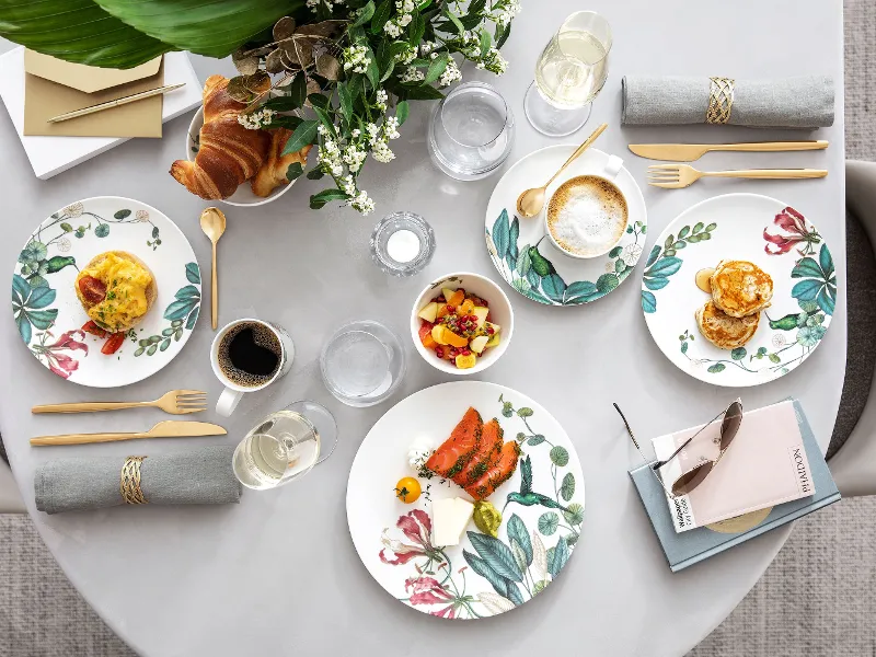 Für eine Mahlzeit gedeckter Tisch mit Blumentellern, Speisen, Getränken, Servietten und einem kleinen Blumenstrauß als Tischdekoration.