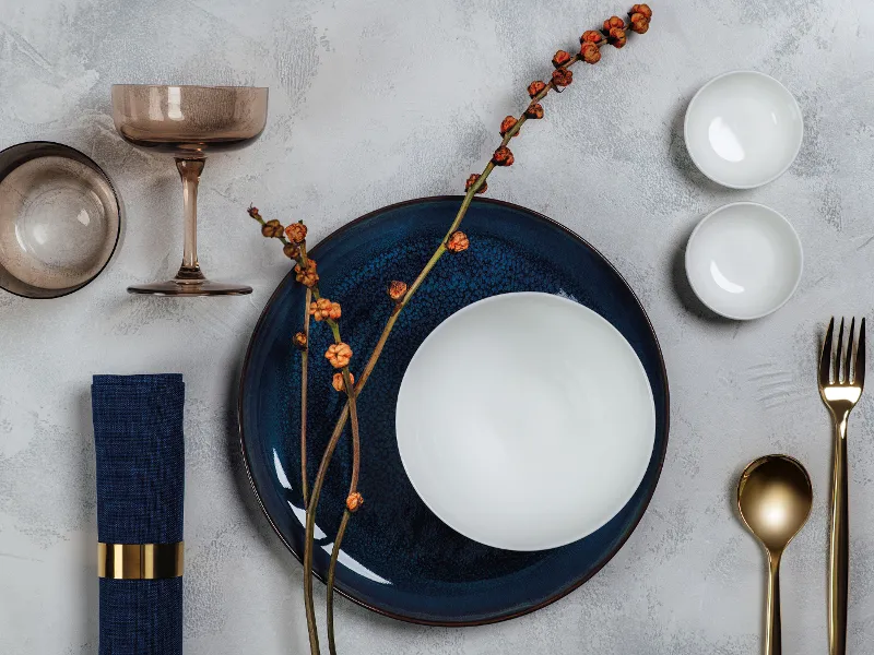 Elegant table setting with a blue Crafted plate, a white Royal bowl, gold cutlery and a dark blue napkin. Delicate dried twigs and two small white bowls on a textured grey surface.