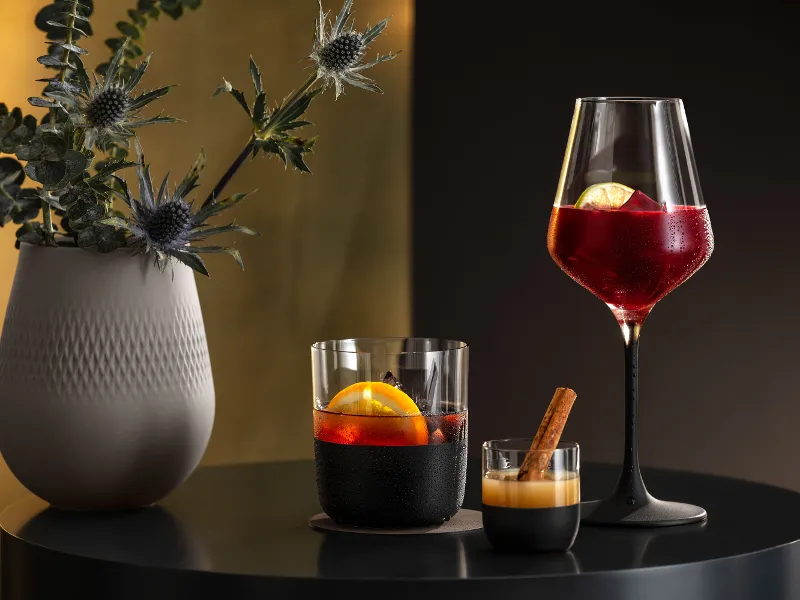 Two cocktails in Manufacture glasses and a Manufacture vase with a plant on a black table against a gold background.