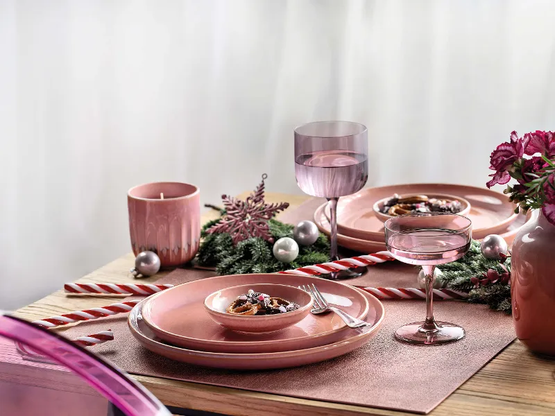 A festive table setting with pink Perlemor crockery, decorated with a snowflake ornament, candy canes, pink flowers and silver baubles. There are also two glasses, a pink candle and a small bowl of food.