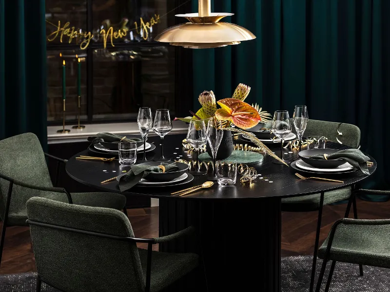 A round black dining table is elegantly set with green napkins, gold cutlery and a flower arrangement. Green chairs surround the table and a "Happy New Year" sign welcomes guests to the New Year's Eve party.