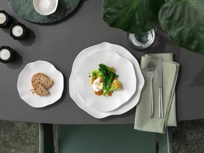 A Pura plate with vegetables and a side dish on a grey table A Pura plate with vegetables and egg, a side plate with bread and cutlery on a napkin on a grey table.