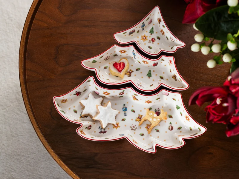 Vista aérea de un juego de 3 cuencos de porcelana para aperitivos con forma de árbol de Navidad de la colección Toy's Delight de Villeroy & Boch sobre una mesa de madera oscura, con galletas de estrellas y un dulce con forma de corazón, al lado de un regalo rojo con lazo de rayas verdes y blancas y flores rojas.