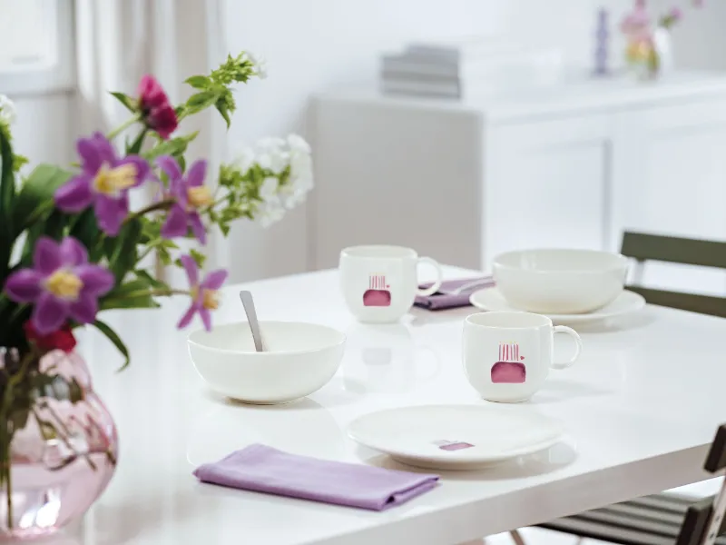 A white table set for two people with bowls, mugs and plates from the With Love collection with a birthday pattern and lavender-coloured napkins. On the left is a vase with purple and white flowers.