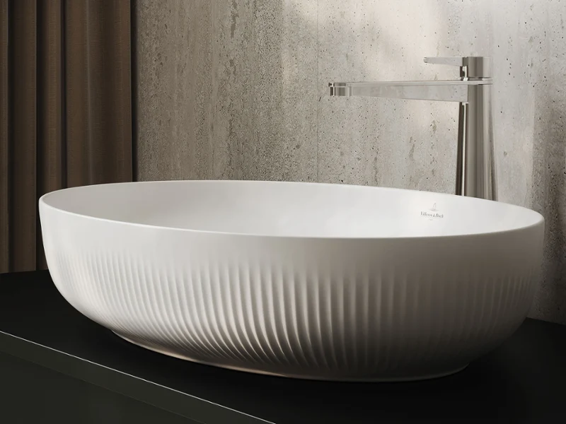 White ceramic washbasin with vertical grooves, placed on a dark countertop in front of a textured wall and a round window.