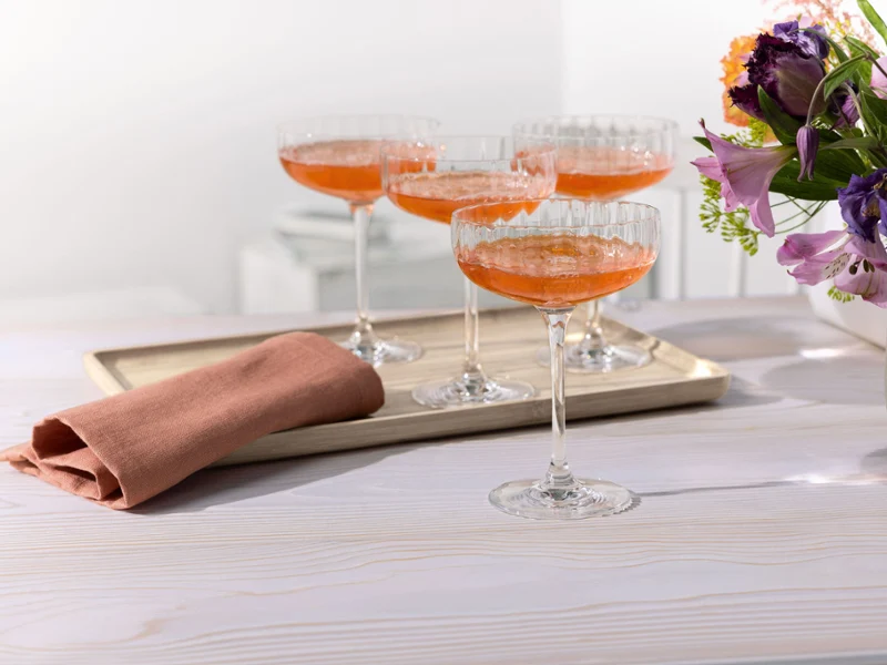 Four Rose Garden coupe glasses with orange cocktails on a tray, next to a folded napkin and a vase of flowers on a white table.