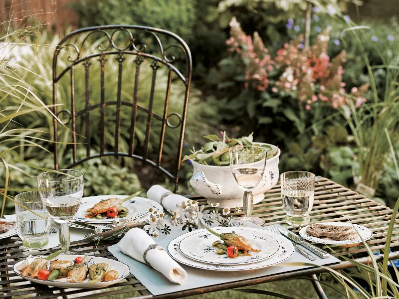 Outdoor dining table setting with three Villeroy & Boch "Old Luxembourg" dinner plates, transparent glasses, a bowl of salad and a wine glass on a striped metal table. A black metal chair and garden plants are visible in the background.