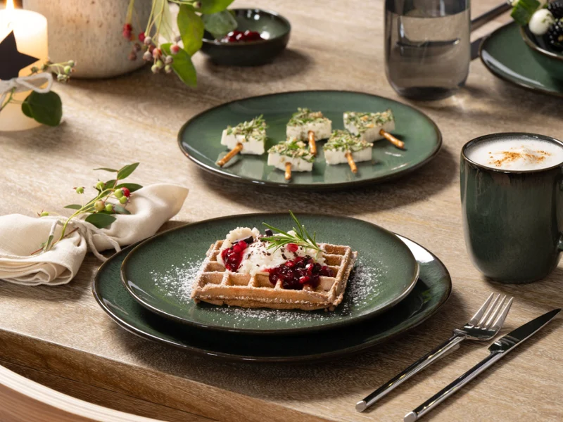 Lave jest idealny na Boże Narodzenie w naturalnych odcieniach Zielony talerz Lave z gofrem z bitą śmietaną i jagodami, kubek kawy Lave i szaszłyki z przekąskami na świątecznie nakrytym drewnianym stole.