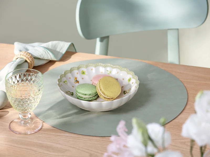 A Easter Delight bowl sits on a light green Easter Delight placemat A Easter Delight bowl of pastel macarons sits on a light green Easter Delight placemat beside a glass and a folded napkin on a wooden table.