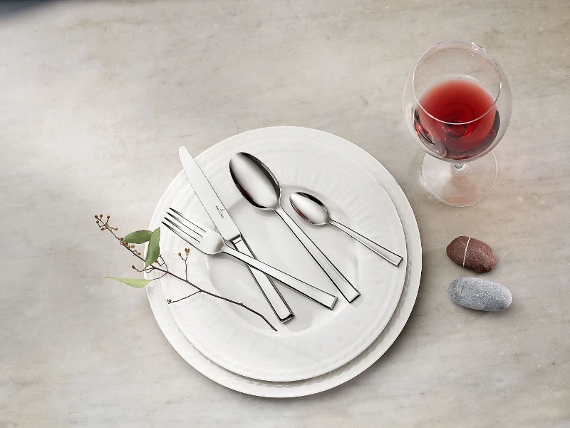 Victor cutlery set on a white Cellini plate A Victor cutlery set with a knife, a fork and two spoons on white Cellini plates, next to a glass of red wine and two smooth stones on a marble surface.