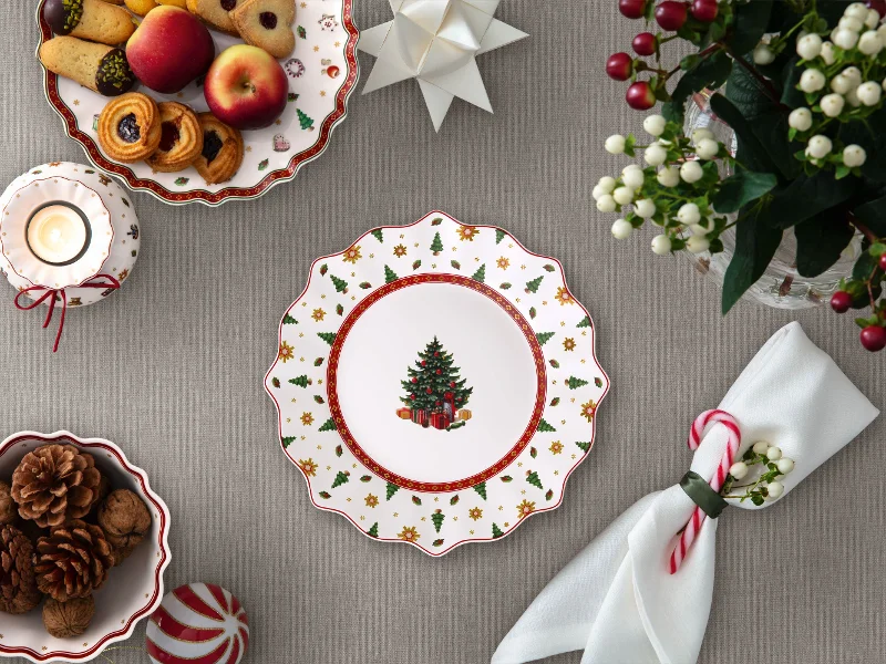 A festively set table with a decorated plate featuring a Christmas tree motif, cookies, fruit, candles, napkins, and a flower arrangement. 
