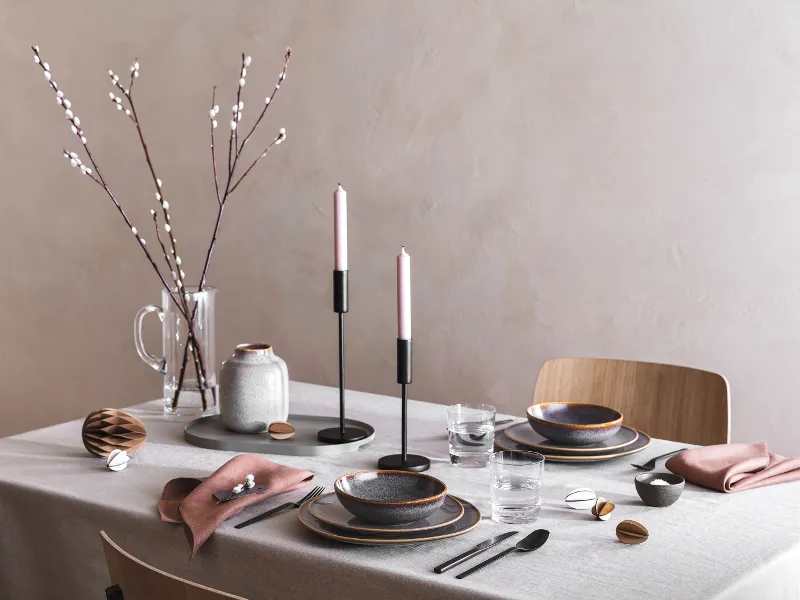 An elegant dining table with lit candles, Lave ceramic crockery, black Manufacture cutlery and minimalist Easter decor with willow catkins.