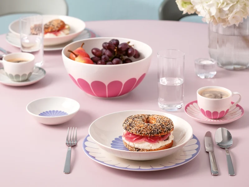 A set breakfast table with a bagel sandwich, a fruit bowl, coffee, water and three place settings on a pink table.
