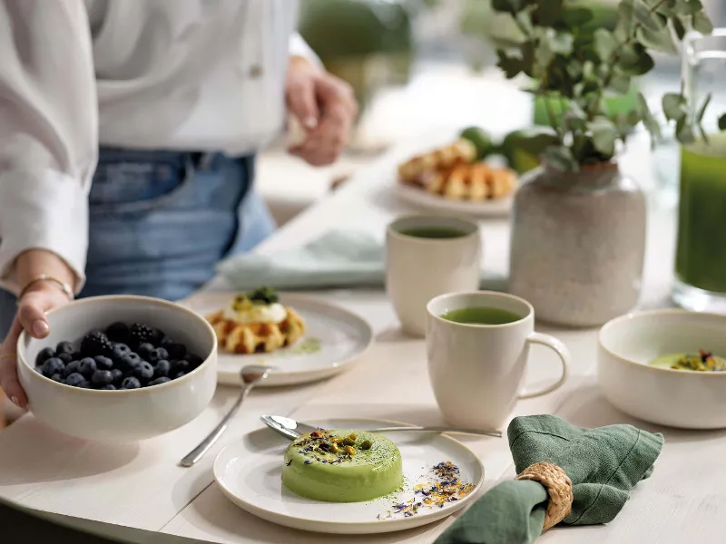 Person preparing a breakfast table with various dishes on Crafted Cotton breakfast plates and with cups of tea.