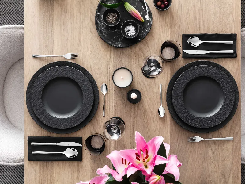 Modern table setting for two with black Manufacture crockery, Piemont cutlery and pink lilies as the centrepiece.