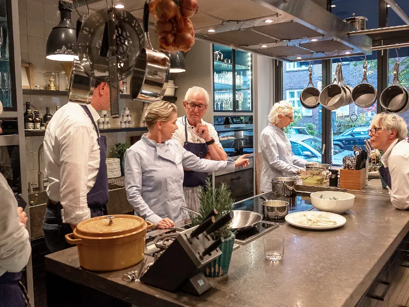 Five people are discussing cooking in a modern kitchen while standing around a counter with pans, plates, and utensils.