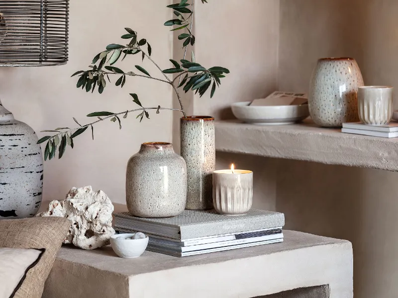 Stylish Lave ceramic vases and a lit candle on top of books, along with a small branch and decorative items on a beige shelf.