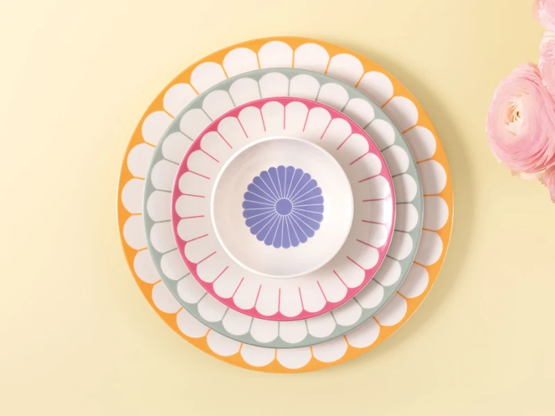 Four Villeroy & Boch Fleur plates in different sizes and colours placed on a light yellow background next to pink flowers.