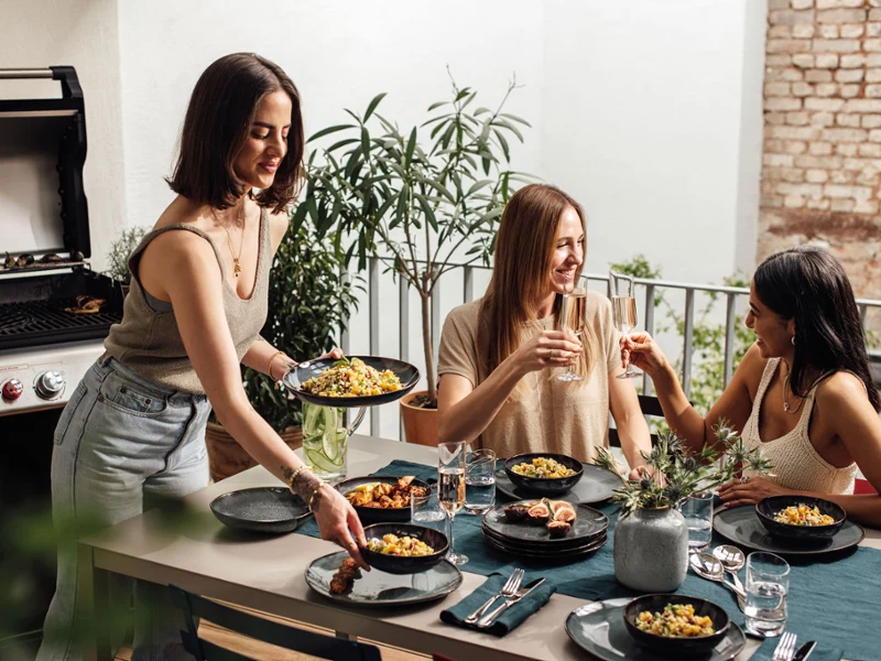 Zwei Freunde genießen sommerliche Leckereien auf Lave Tellern und stoßen mit einem Glas Sekt an, eine weitere Person serviert Essen.