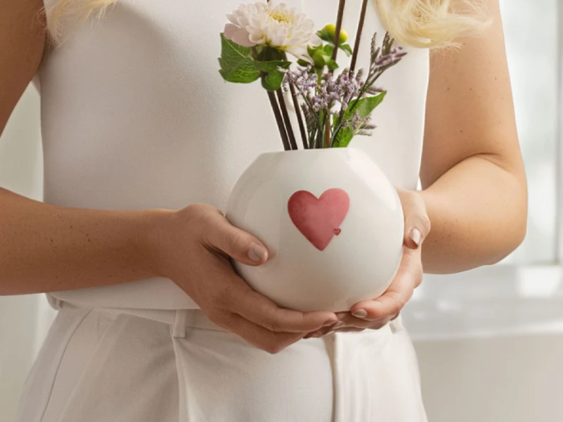 Okrągły wazon marki with love z czerwonym motywem serca Kobieta trzymająca okrągły biały wazon marki with love z czerwono-różowym sercem i białymi kwiatami.