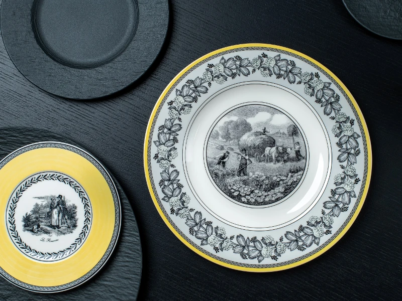 Elegant plates on a dark surface; one large with a rural scene and floral border, another smaller with a yellow rim depicting a couple.